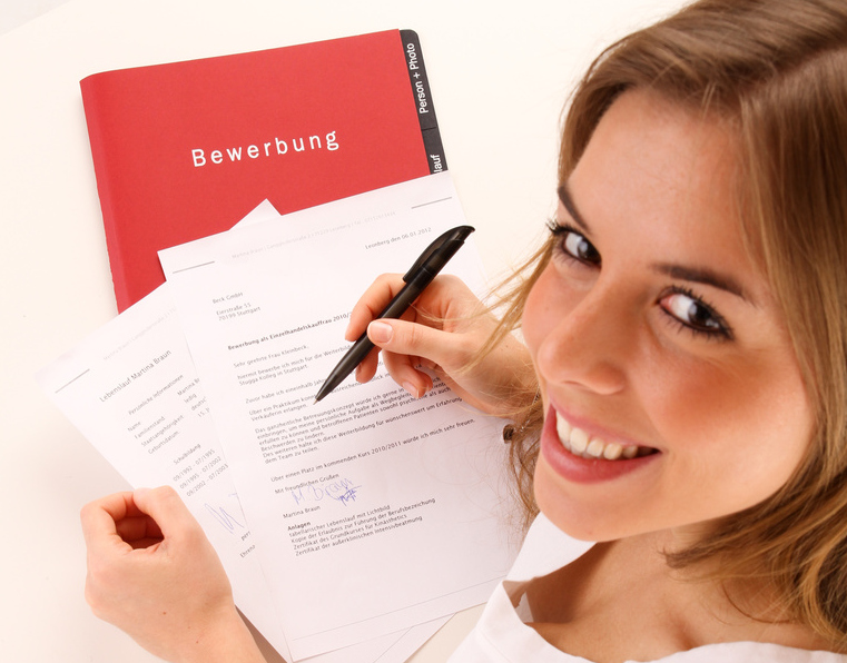 Eine junge Frau schreibt an ihre Bewerbung, eine rote Bewerbungsmappe liegt auf dem Tisch